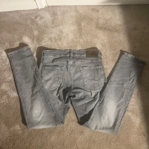Grå jeans från Jack & Jones - Snygga grå jeans från Jack & Jones i modellen Slim Glenn. De har en slim passform och klassisk femficksdesign med knapp och dragkedja. Perfekta för en stilren look.