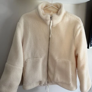 Beige teddyjacka med dragkedja - Mysig beige teddyjacka med dragkedja framtill och två stora fickor. Perfekt för kyliga dagar när du vill hålla dig varm och bekväm. Jackan har en hög krage och en avslappnad passform. Aldrig använd. 