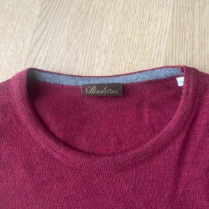 Vinröd tröja från Stenströms - Säljer en stilren vinröd tröja från Stenströms med grå armbågslappar. Tröjan har rund hals och långa ärmar, perfekt för kyligare dagar. Den är tillverkad i ett mjukt material som ger en bekväm passform. Den har ett hål vid bröstet(se sista bilden) annars är den i topp skick 