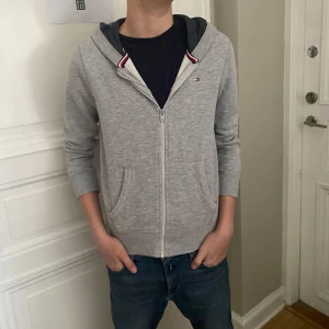 Grå zip hoodie från Tommy Hilfiger - Säljer en stilren grå hoodie från Tommy Hilfiger. Hoodien är i bra skick och perfekt nu på våren och till sommarkvällar. Säljer tröjan då den är för liten på mig. Modellen på bilden är 166cm lång och väger 45kg. Passar förmodligen bäst om du är mellan 155-160. (Nypris ca 1000kr) mitt pris 349 kr