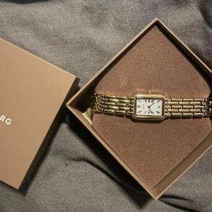 Guldfärgad klocka med vit urtavla - Elegant guldfärgad klocka med rektangulär boett och vit urtavla. Klockan har romerska siffror och en länkarmband i guld. Perfekt som en stilren accessoar.