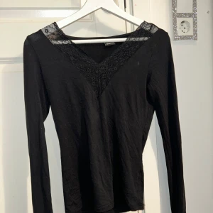 Svart långärmad topp från Gina Tricot - Snygg svart långärmad topp från Gina Tricot med spetsdetaljer vid halsringningen. Perfekt för en stilren look. Toppen har en v-ringad design och är tillverkad i ett mjukt material.