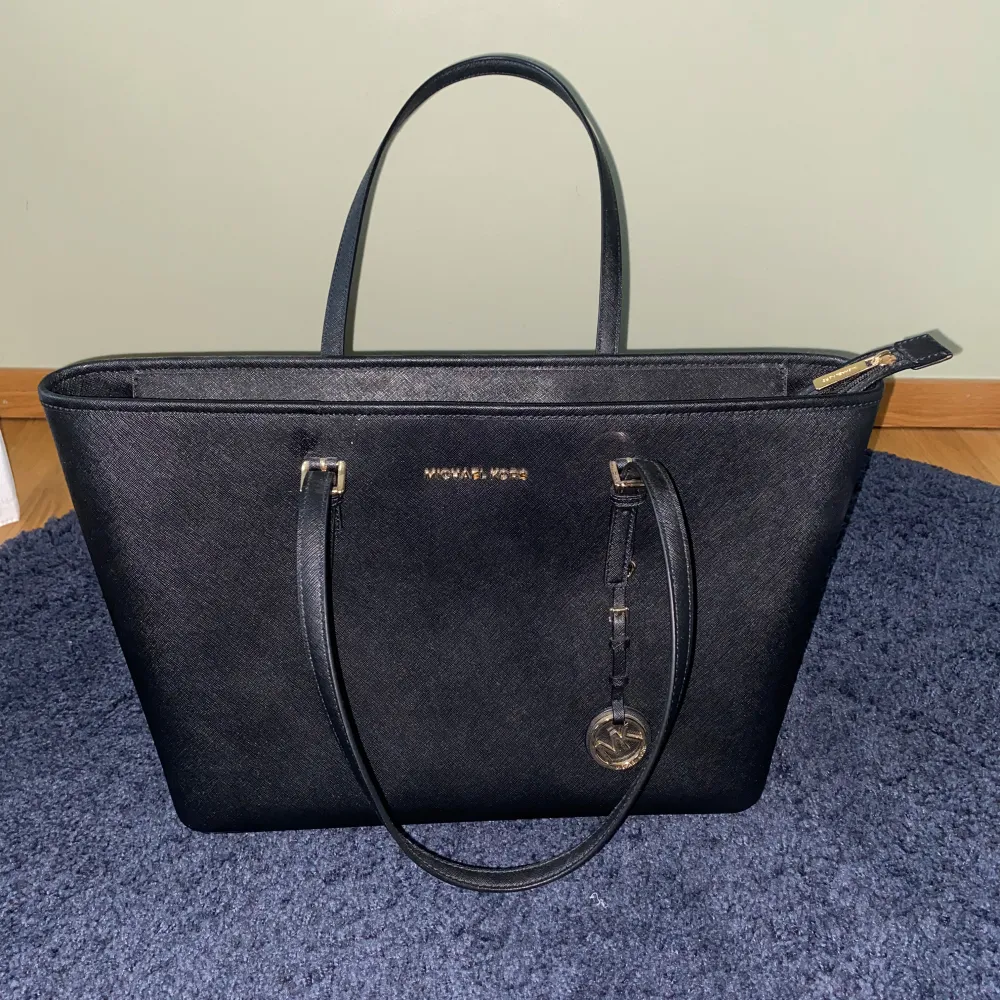 Snygg svart axelväska från Michael Kors med guldfärgade detaljer och logotyp. Väskan har en dragkedja upptill och flera fack inuti för smart förvaring. Perfekt för att bära med sig det viktigaste med stil. Väskan har dessutom fack för dator. Den är oanvänd och förpackningen ingår (sista bilden). Laukut & Käsilaukut.