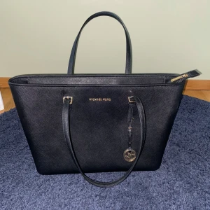 Svart axelväska från Michael Kors - Snygg svart axelväska från Michael Kors med guldfärgade detaljer och logotyp. Väskan har en dragkedja upptill och flera fack inuti för smart förvaring. Perfekt för att bära med sig det viktigaste med stil. Väskan har dessutom fack för dator. Den är oanvänd och förpackningen ingår (sista bilden)