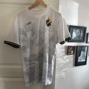Vit AIK fotbollströja från Nike - Säljer en vit AIK fotbollströja från Nike med grått mönster och klubbens emblem på bröstet. Tröjan har korta ärmar med svarta och gula detaljer. Perfekt för AIK-fans! 🖤💛