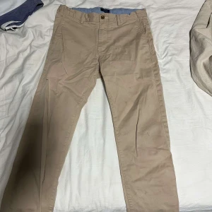 Beige chinos från GANT - Säljer ett par stilrena beige chinos från GANT. Byxorna har en klassisk design med knappar och fickor både fram och bak. Perfekta för en avslappnad men ändå uppklädd look. Finns få fläckar