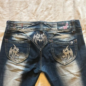 Blå jeans från Crazy Age - Säljer jeans från Crazy Age med coola drakmotiv på bakfickorna, då de inte är min stil längre. Jeansen är bootcut och det står att de är large men jag skulle säga att de passar mer som en small. Väldigt lågmidjade och har smickrande passform. Använda endast ett fåtal gånger och har inga defekter förutom att ett par rhinestones ramlat av