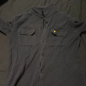 Mörkblå skjorta från Lyle & Scott - Snygg mörkblå skjorta från Lyle & Scott med klassisk krage och knäppning fram. Skjortan har två bröstfickor med knappar och en gul broderad logotyp på ena fickan. Perfekt för en stilren look.