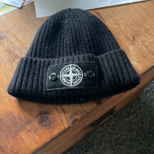 Svart ribbad mössa från Stone Island - Snygg svart ribbad mössa från Stone Island med det ikoniska märket framtill. Perfekt för att hålla värmen under kyliga dagar. Stilren och enkel att matcha med olika outfits. Med vitt märke 
