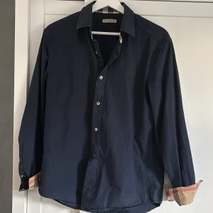 Mörkblå skjorta från Burberry Brit - Snygg mörkblå skjorta från Burberry Brit med klassiska rutor på insidan av kragen och ärmsluten. Skjortan har långa ärmar och knappar framtill. Perfekt för en stilren look.
