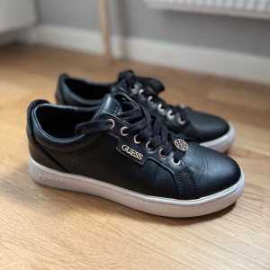 Svarta sneakers från Guess - Svarta sneakers från Guess med vit sula och gulddetaljer. Perfekt nu till våren och sommaren. Endast använda ett fåtal gånger.  Storlek 38.