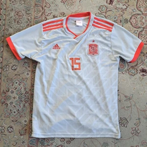 Spanien fotbollströja - Säljer en vit fotbollströja från det Spanska landslaget med röda detaljer och nummer 15. Tröjan har korta ärmar och ett tryck med 'Ramos' på baksidan. Perfekt för fotbollsfans!
