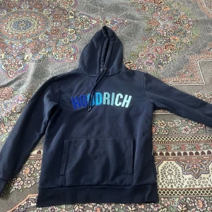 Svart hoodie från Hoodrich - Snygg svart hoodie från Hoodrich med blå och vit text på bröstet. Den har en stor ficka framtill och justerbar huva med snören. Perfekt för en avslappnad stil.