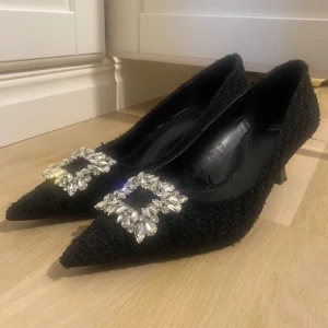Svarta klackskor i tweed med strass - Eleganta svarta pumps med spetsig tå och glittrig spänne framtill. Skorna är i tweed och en liten klack som ger en sofistikerad look. Endast använda en gång.