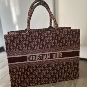 Mönstrad shoppingväska från Christian Dior - Snygg shoppingväska från Christian Dior med ett ikoniskt mönster i rött och beige. Väskan har två handtag och är rymlig nog för alla dina dagliga behov. Perfekt för en stilfull shoppingtur eller som en elegant accessoar.
