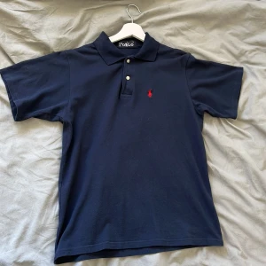 Mörkblå pikétröja från Polo - Säljer en klassisk mörkblå pikétröja från Polo med korta ärmar och en liten röd logga på bröstet. Tröjan har en krage med två knappar och är perfekt för en stilren look. Storlek Small
