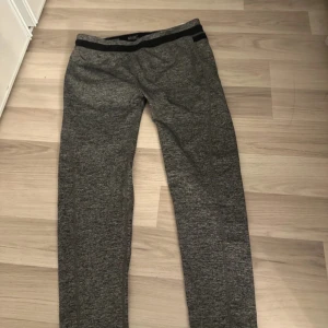 Grå WR.UP byxor med svart midjeband - Snygga grå byxor från WR.UP med svart elastiskt midjeband. De har en slim passform och är tillverkade i ett mjukt syntetmaterial som sitter skönt mot kroppen. Perfekta för träning eller chill hemma. Byxorna har en modern look med diskreta sömmar och en sportig vibe.