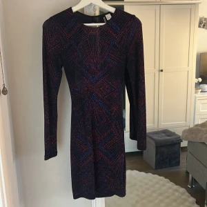 Svart glittrig kortklänning Divided - Säljer en svart kortklänning från Divided med glittrigt geometriskt mönster i rött och blått. Klänningen har långärmade ärmar och en snygg öppen rygg som ger extra edge. Perfekt för dig som vill sticka ut med coola detaljer och glans.