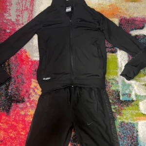 Svart Nike träningsset med dragkedja - Nike träningsset i helsvart med diskret logga på både jacka och byxa. Jackan har dragkedja framtill och höga muddar, byxorna har snörning i midjan och sidopaneler i blankare material. Perfekt för dig som gillar sportig och clean stil.