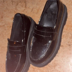 Svarta chunky loafers med platt sula - Säljer ett par svarta loafers med chunky platt sula och rund tå. Skorna har en glansig finish och är tillverkade i syntetmaterial. Perfekt för dig som gillar en stilren och edgy look. Passar till både jeans och kostymbyxor.