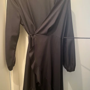 Svart omlottklänning med volang - Det Snygg svart omlottklänning med långa ärmar och volangdetalj längs kanten. Klänningen har en elegant v-ringning och knyt i midjan för en smickrande siluett. Perfekt för dig som gillar stilrena och trendiga plagg.