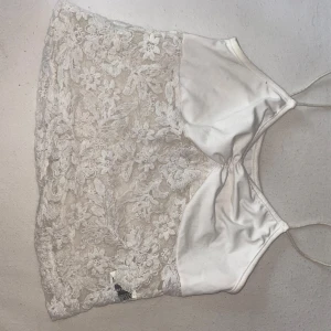 Vit spetsig linnetopp med satin - Supersöt vit linnetopp med tunna axelband. Toppen har en snygg kombination av glansig satin framtill och blommig spets över hela ryggen och sidan. 