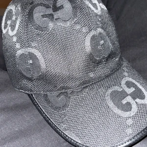 Svart keps från Gucci med GG-mönster - Snygg svart keps från Gucci med det klassiska GG-mönstret över hela kepsen. Materialet är slitstark polyester med en lätt glansig finish. Perfekt för dig som vill ha en lyxig och trendig accessoar med tydlig designerstil.