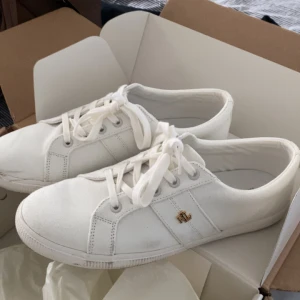 Vita sneakers från Ralph Lauren - Snygga vita sneakers från Ralph Lauren med klassisk låg profil och rund tå. Skorna har snörning och är tillverkade i bomullstyg med en liten guldfärgad RL-logga på sidan. Perfekta för dig som gillar stilrena och fräscha sneakers.