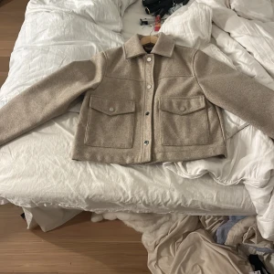 Beige overshirt med stora fickor - Snygg beige overshirt med klassisk krage och stora fickor framtill. Jackan har knappar hela vägen och är tillverkad i ett mjukt ullmaterial. Perfekt för lager-på-lager och har en clean, modern look som funkar till jeans eller dressade byxor.