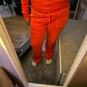 Orange mjukisbyxor från Juicy Couture - Säljer ett par riktigt snygga orange mjukisbyxor från Juicy Couture i mjuk sammet. Byxorna har bred resår i midjan med snörning, stora fickor fram och bak med silverfärgade detaljer samt broderad logga på ena fickan. Perfekt för chill eller streetwear.