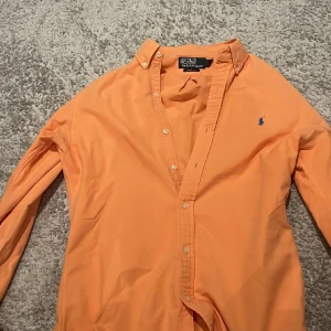 Orange skjorta från Polo Ralph Lauren - Slim fit orange skjorta från Polo Ralph Lauren med klassisk krage och blå broderad logga på bröstet. Skjortan har långa ärmar och knappar framtill. Perfekt för dig som gillar stilrena och färgstarka plagg.