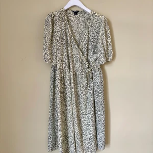 Blommig omlottklänning från Monki - Säljer en ljus omlottklänning från Monki med svart blommigt mönster. Klänningen har v-ringning, puffiga korta ärmar och knyt i midjan. Längden är mitt på vaden och tyget är mjukt och luftigt, perfekt för varma dagar.