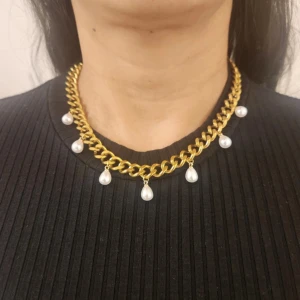 Guldfärgat halsband med pärldroppar - Chunky guldfärgat halsband med stora länkar och hängande vita pärldroppar. Statement-smycke som ger en lyxig känsla och passar perfekt när du vill sticka ut. Pärlorna är droppformade och sitter jämnt fördelade längs hela halsbandet.