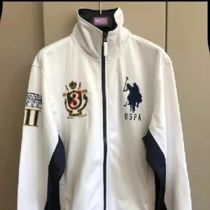 Vit jacka/tröja  från US Polo Assn - Snygg vit jacka/tröja  från US Polo Assn med marinblå detaljer och broderade emblem på bröstet och ärmen. Jackan har hög krage, dragkedja framtill och sportig look. Perfekt för dig som gillar klassisk stil med en twist. USPA-logga och nummerdetaljer ger extra karaktär.