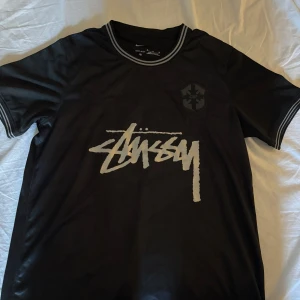 Svart Stüssy x Nike fotbollströja M - Snygg svart fotbollströja från Nike och Stüssy i Dri-FIT material. Tröjan har korta ärmar, vit Stüssy-logga på bröstet och diskret CBF-märke. Ribbade detaljer i vitt på krage och ärmslut ger en clean look. Perfekt för dig som gillar streetwear och fotboll. Otroligt bra skick 10/10
