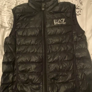 Svart dunväst från EA7 Armani - Svart dunväst från EA7 Emporio Armani med quiltad design och hög krage. Västen har en tydlig logga på bröstet och stängs med dragkedja framtill. Perfekt för lager-på-lager och har en clean, sportig look som funkar till många stilar.
