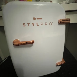 STYLPRO sminkkylskåp i vitt och roséguld - Snyggt sminkkylskåp från LYKO i vit plast med detaljer i roséguld. Perfekt för att förvara dina favoritprodukter som ansiktsmist, serum och sheet masks. Kompakt design med hylla och blå insida, passar på sminkbordet eller i badrummet.