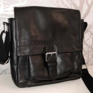 Marc O’Polo svart axelväska i skinn - Snygg svart axelväska från Marc O’Polo i skinn med justerbar rem och praktiskt spänne framtill. Väskan har en fyrkantig form, ett stort huvudfack, en ficka med dragkedja på baksidan och en innerficka. 