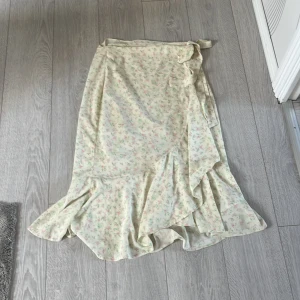 Blommig omlottkjol från Gina Tricot - Supersöt lång omlottkjol från Gina Tricot i ljusbeige med små rosa och gröna blommor. Kjolen har volangkant och knyt i midjan för en extra feminin vibe. Perfekt för dig som gillar romantiska och somriga looks!