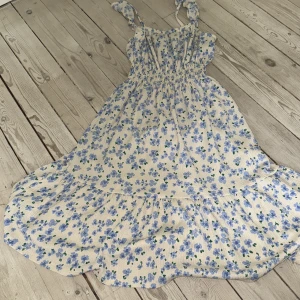 Blommig sommarklänning från Zara - Supersöt långklänning från Zara med ljusbeige botten och blå blommor. Klänningen har smock i midjan, volang nedtill och breda axelband med knyt. Perfekt för varma dagar och har en flowy, avslappnad vibe. Materialet känns mjukt och luftigt.