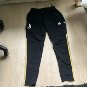 Juventus svarta träningsbyxor Adidas - Svarta träningsbyxor från Adidas med Juventus-logga på ena benet och vita Adidas-märket på andra. Snygga gula ränder längs sidorna och reflekterande detaljer baktill. Byxorna har smal passform och är perfekta för träning eller chill. Elastisk midja med snörning för skön passform.