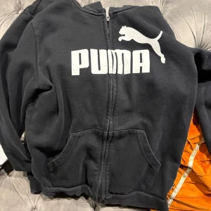 Svart hoodie från Puma  - Svart hoodie från Puma i storlek 164 (13 till 14 år)med stor vit logga och puma-tryck på bröstet. Tröjan har dragkedja framtill, två fickor och en skön huva. Materialet är mjuk bomull och polyester, perfekt för chill eller träning. Klassisk streetwear-look.