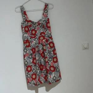 Blommig kortklänning från Fröken Söt - Säljer en söt kortklänning från Fröken Söt med breda axelband och blommigt mönster i rött, rosa, beige och grått. Klänningen har smockad rygg och är tillverkad i mjuk bomull, perfekt för varma dagar med underkjol. Passformen är normal och modellen är luftig och bekväm.