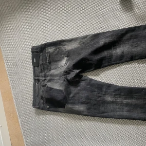 Svarta slitna jeans från Replay - Säljer ett par svarta jeans från Replay med coola slitningar och hål på benen. Jeansen har en skinny passform och klassisk femficksdesign. Färgen är mörk med blekta detaljer och materialet är stretchigt denim för skön känsla. Perfekt för dig som gillar en edgy streetstil.