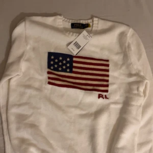 Vit stickad tröja Polo Ralph Lauren - Vit stickad tröja från Polo Ralph Lauren med amerikansk flagga i blått och rött på bröstet samt RL-broderi. Rund hals och ribbade muddar vid ärmslut och nederkant. Perfekt för dig som gillar klassisk och clean stil med en touch av USA. Pris 999
