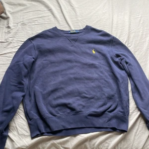 Marinblå sweatshirt från Polo Ralph Lauren - Säljer en marinblå sweatshirt från Polo Ralph Lauren i storlek M. Tröjan har klassisk rund hals, långärmad design och gul broderad logga på bröstet. Mjuk bomullskvalitet och ribbade muddar vid ärmslut och nederkant. Perfekt för chill dagar.