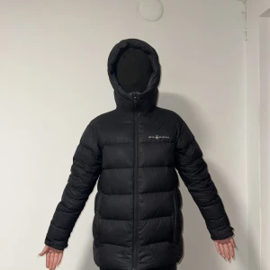 Svart dunjacka från Sail Racing - Svart dunjacka från Sail Racing med huva och dragkedja framtill. Jackan är quiltad och har en clean look med logga på bröstet och bak på huvan. Fylld med Allied Feather Down 600 för extra värme. Perfekt för kalla dagar och har en schysst oversized passform.