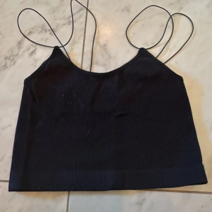 Svart ribbad linnetopp från Bik Bok - Svart ribbad linnetopp från Bik Bok med tunna axelband och croppad passform. Materialet är mjukt och stretchigt med 92% polyamid och 8% elastan. Perfekt för dig som gillar minimalistisk stil och vill ha en enkel topp med snygg struktur.