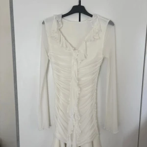 Vit omlottklänning med volanger från SHEIN - Supersöt vit kortklänning från SHEIN i storlek XS. Klänningen har långärmade meshärmar, omlott framtill med volangdetaljer och snygg rynkning längs hela kroppen. Kjolen är lätt utsvängd med volangkant för extra flow. Perfekt för dig som gillar romantisk och trendig stil.