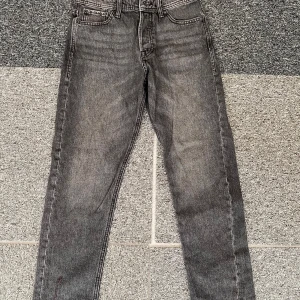 Grå loose fit jeans Jack & Jones - Säljer ett par grå jeans från Jack & Jones, modell Loose/Chris. De har en loose fit med raka ben och klassisk femficksdesign. Jeansen är tillverkade i denim och har svarta detaljer vid knappar och nitar. Perfekt för dig som gillar avslappnad stil.